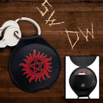 A round, black coin holder with two keys on a key ring at the top. In the center is a red embossed image of a the anti-possession symbol. Behind the holder is a wooden table with the initials SW and DW carved into it. At the bottom right is an inset image of holder opened up with a Challenge Coin inside.