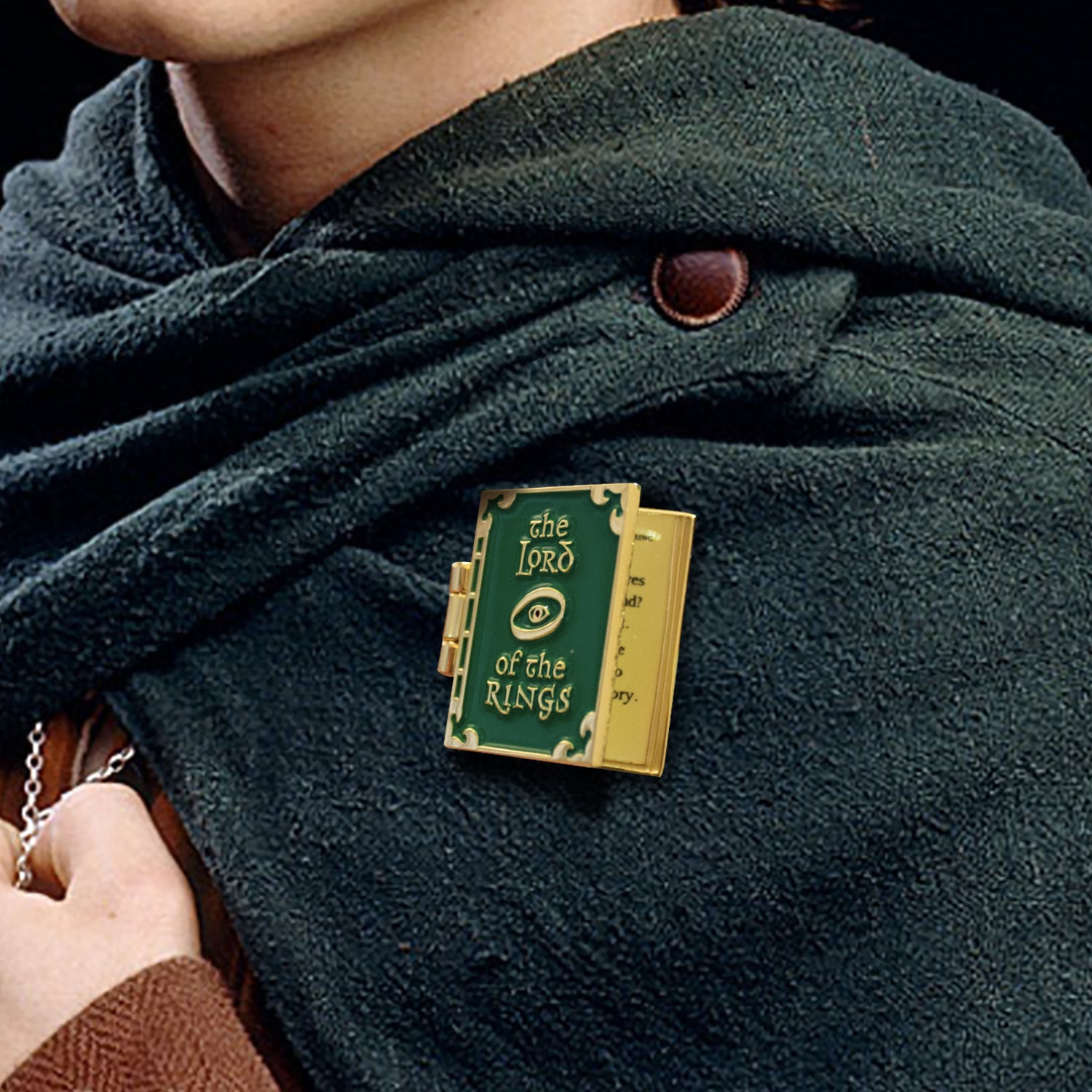A book-shaped enamel pin, partially opened like a book, fastened to a green cloak. The front of the pin is green, with "The lord of the rings" and an image of the eye of sauron in gold. The interior of the pin is gold plated, with a quote from The Lord Of The Rings in black text.