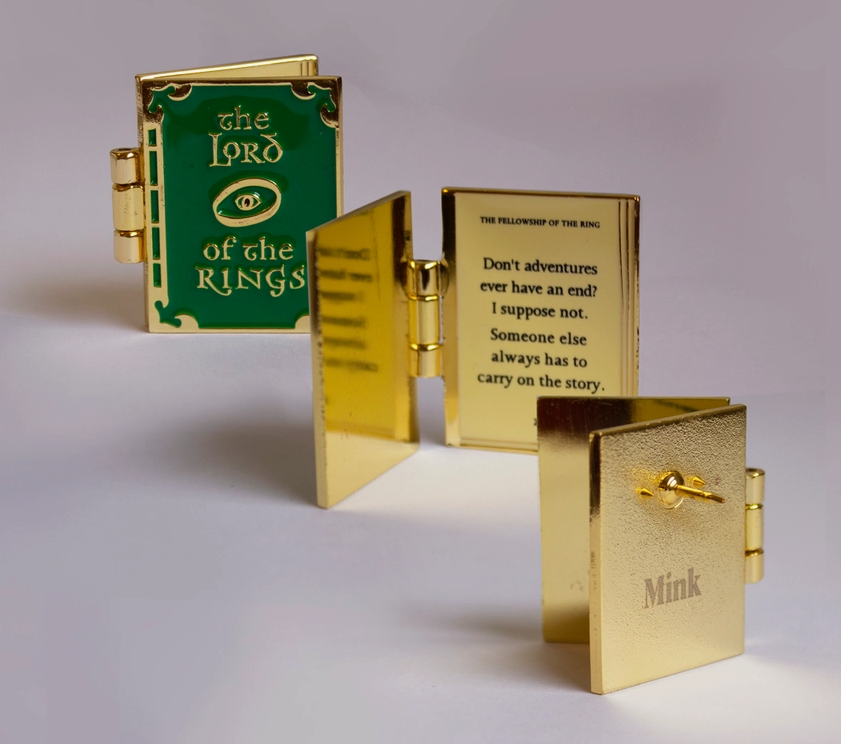 Three views of a hinged enamel pin in the shape of a tiny book. The front of the pin is green, with "The lord of the rings" and an image of the eye of sauron in gold. The back of the pin is gold with a pin post sticking out. The center view shows the pin spread open like a book, with a quote from the lord of the rings in black text.
