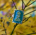 A book-shaped enamel pin sitting on a leafy twig. The front of the pin is green, with "The lord of the rings" and an image of the eye of sauron in gold.
