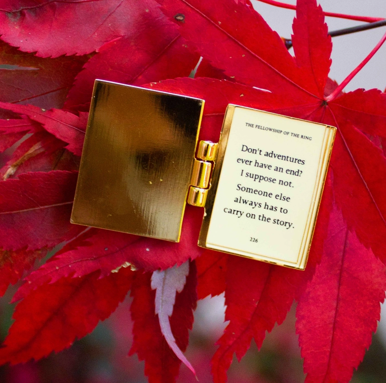 A rectangular enamel pin, opened like a book, on a read leaf. The interior of the pin is gold plated, with a quote from The Lord Of The Rings in black text.