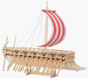 Side view of a miniature wooden Viking ship with a read and white sail, set against a white background. The ship has many oars sticking out the sides, and miniature wooden shields line the edge, with drawings of axes and dragon heads on each.