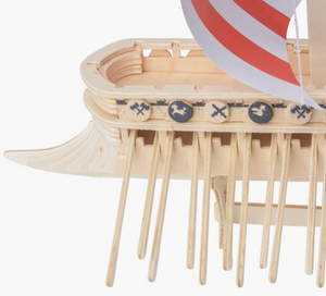 Close up view of the aft section of a miniature wooden Viking ship with a read and white sail, set against a white background. The ship has many oars sticking out the sides, and miniature wooden shields line the edge, with drawings of axes and dragon heads on each.