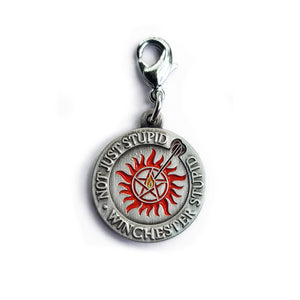 A flat circular charm that says "Not just stupid, Winchester stupid" around the edge, with an anti-possession symbol and a flaming arrow shot into the middle of it. Art by Charlotte Hill.